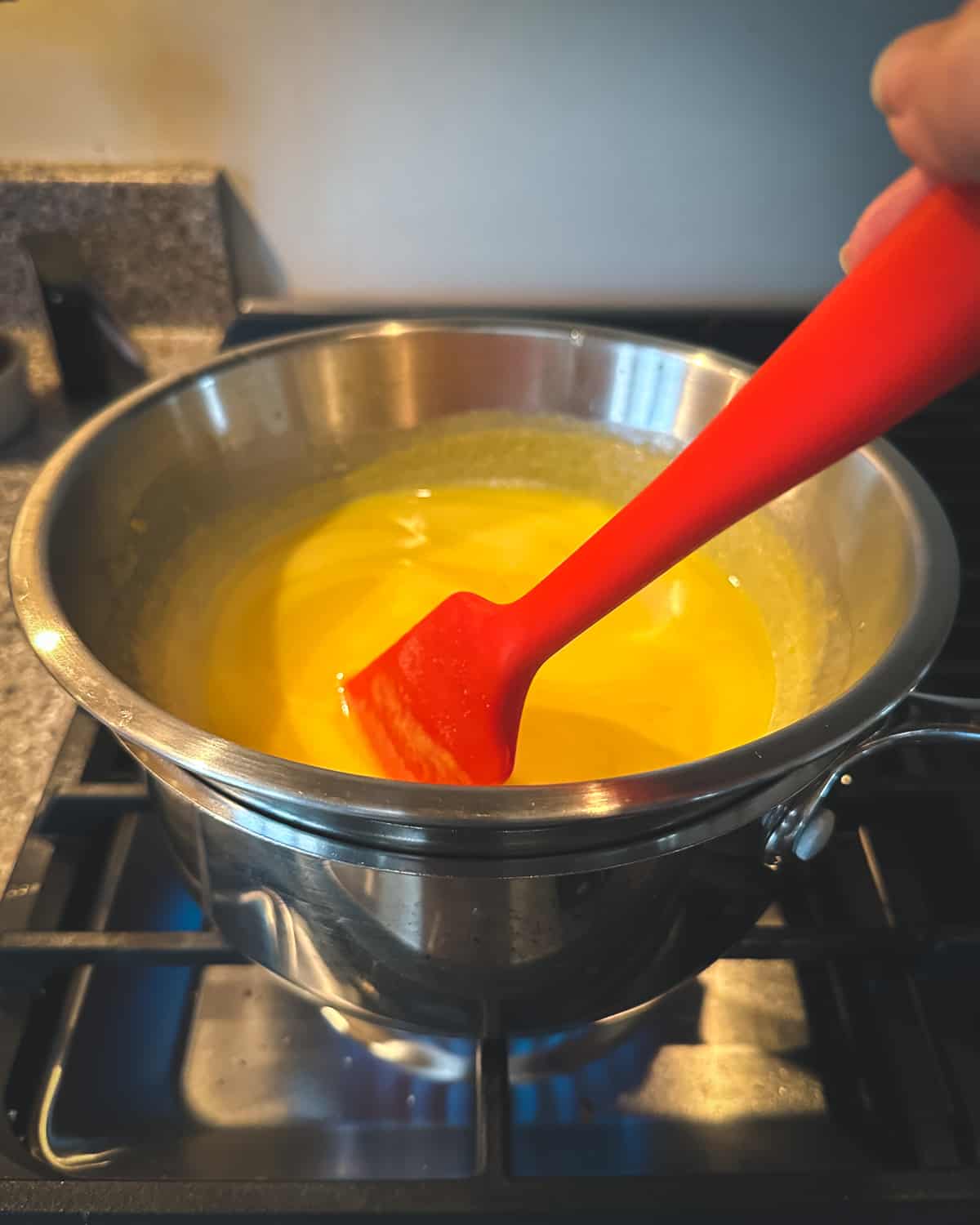 A makeshift double boiler with a metal bowl, and a red spatula stirring the orange curd as it warms. 