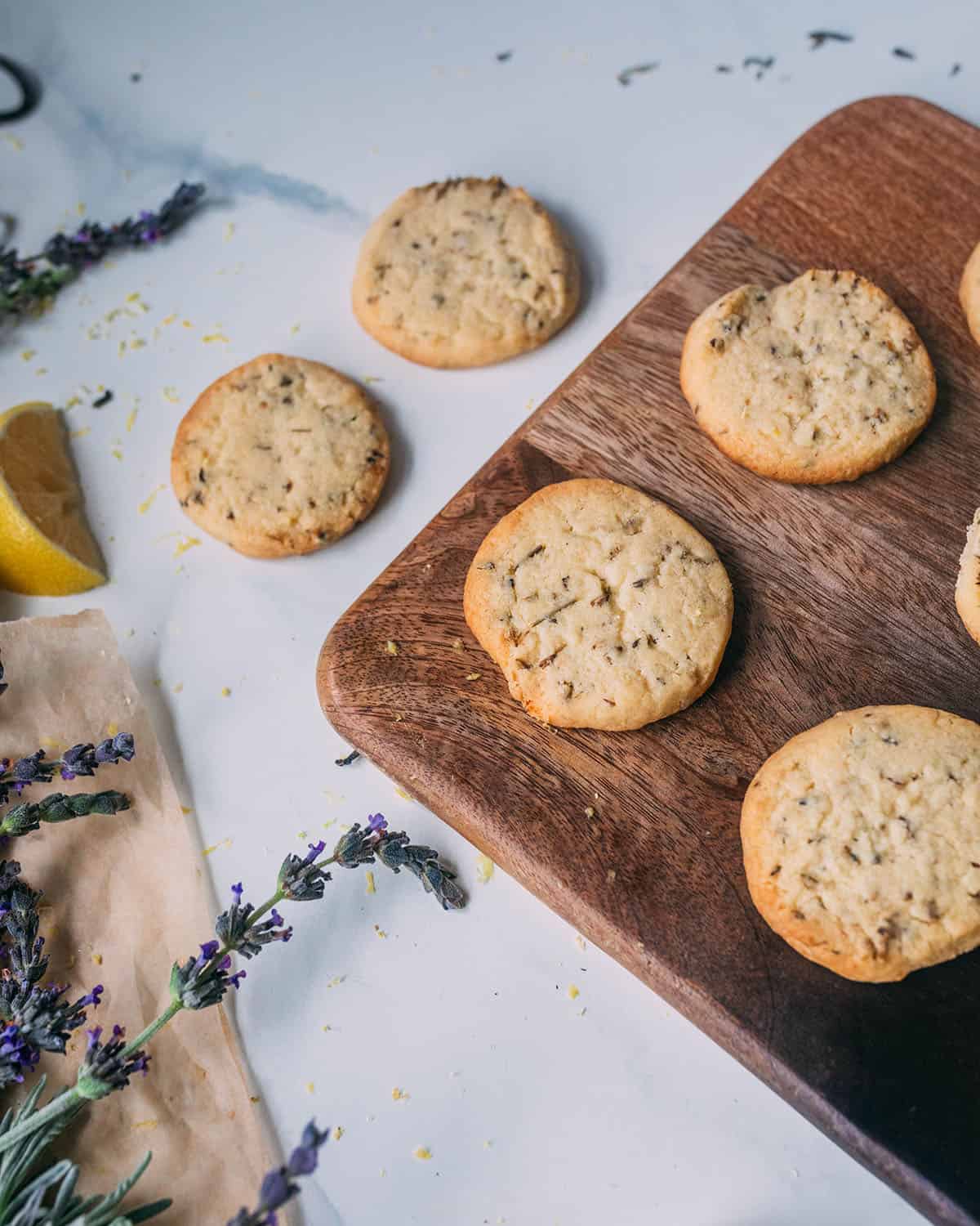 Lemon lavender cookies on a wooden cutting board and a white countertop, showing the corner of the cutting board. Surrounded by fresh lavender and lemon slices.