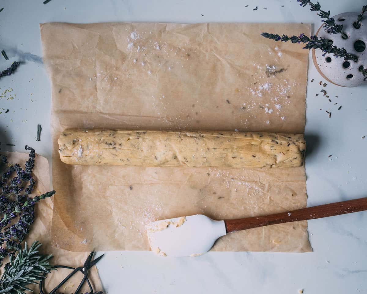 Lavender lemon shortbread cookie dough formed into a log resting on brown parchment paper with a white spatula laying next to it, surrounded by fresh lavender.