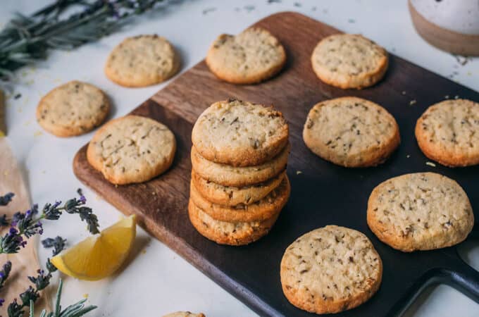 A stack of lemon lavender shortbread cookies on a dark wooden cutting board with a light background, surrounded by other lemon lavender shortbread cookies, and fresh lavender.