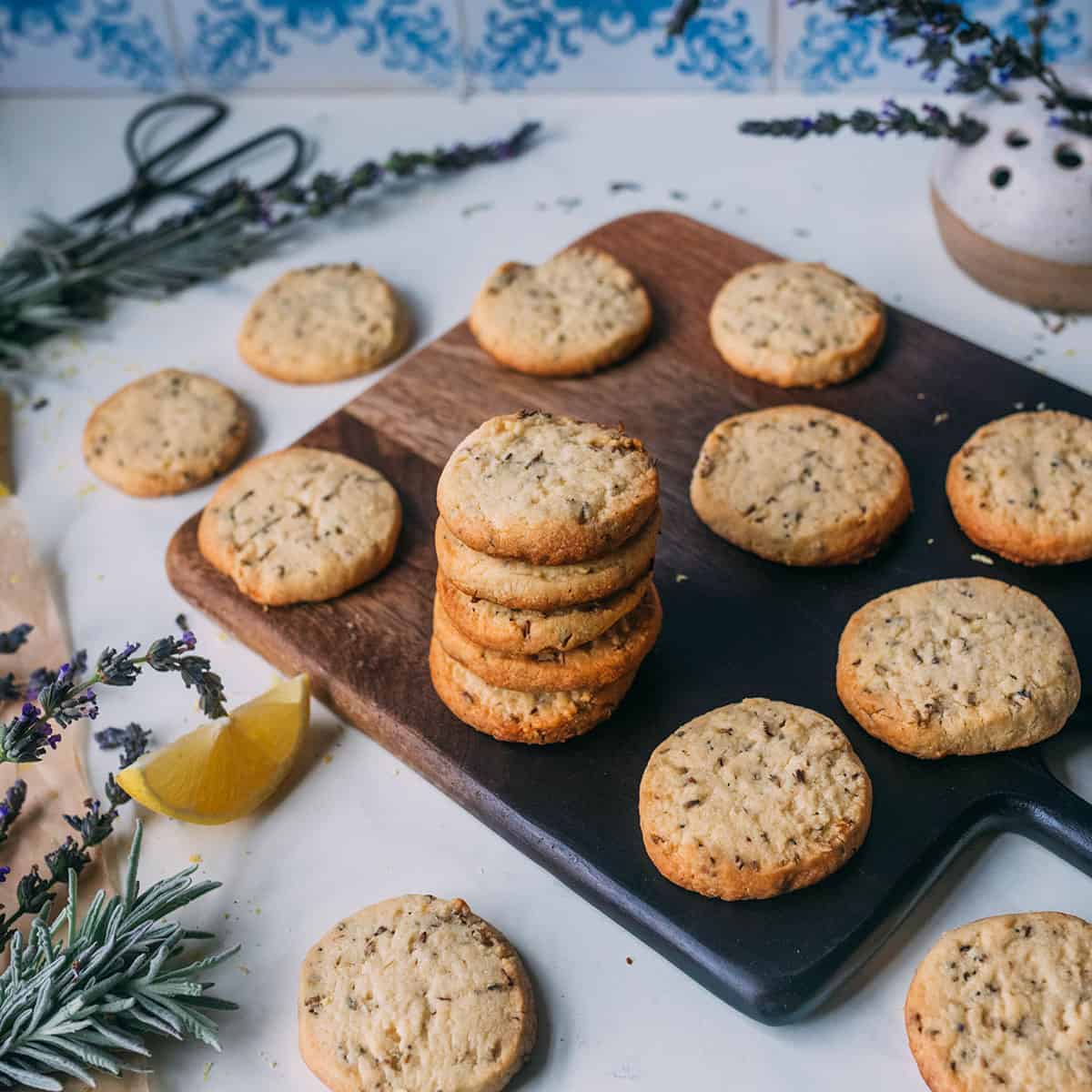 A stack of lemon lavender shortbread cookies on a dark wooden cutting board with a light background, surrounded by other lemon lavender shortbread cookies, and fresh lavender. 