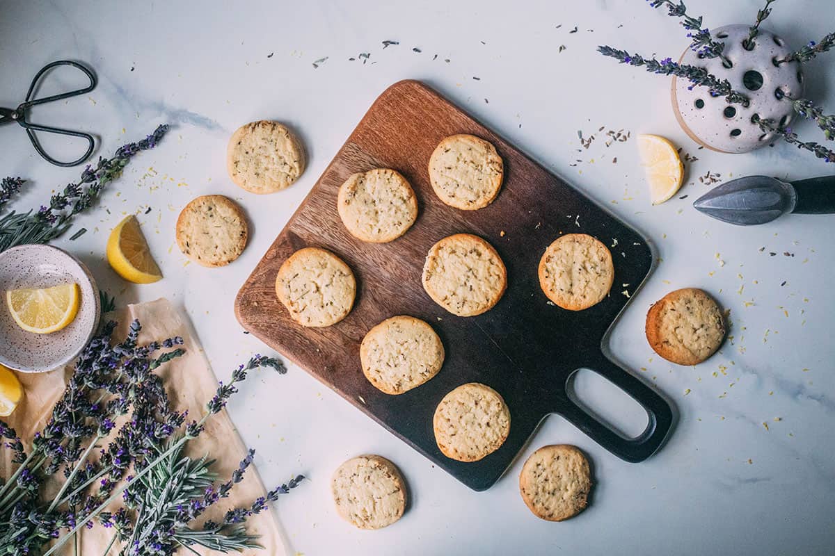 Lemon lavender shortbread cookies on a dark wooden cutting board on a white surface, with cookies also on the countertop. Surrounded by fresh lavender and lemon slices.