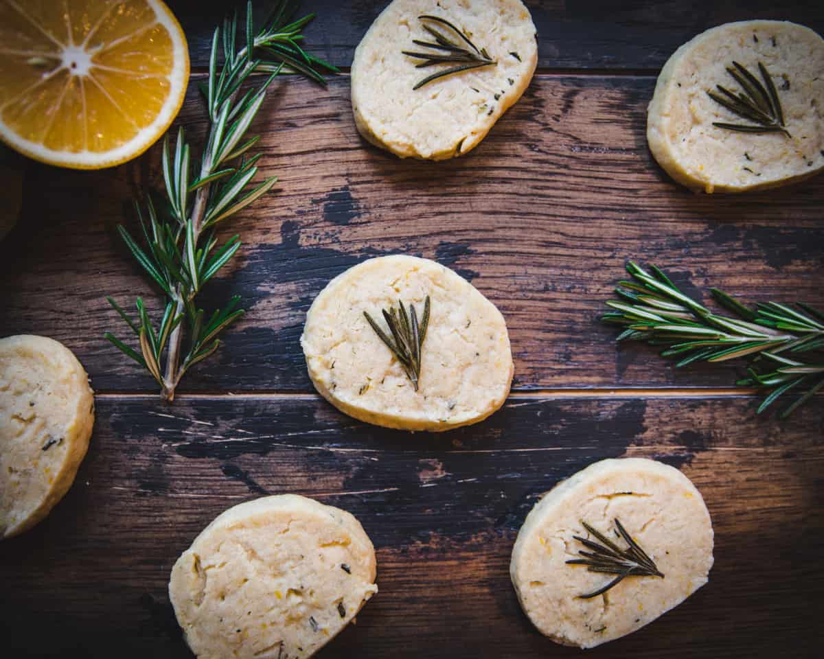 Lemon shortbread cookies with rosemary sprigs on top, surrounded by fresh rosemary and lemon halves on a dark wood surface. 