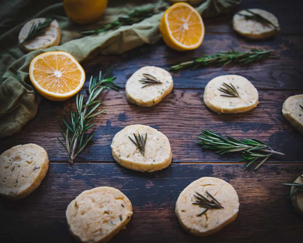 Lemon shortbread cookies on a dark wood surface with fresh rosemary twigs, halved lemons, and green fabric surrounding.