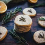 A stack of lemon rosemary shortbread cookies surrounded by single cookies each with a fresh rosemary frond on top, on a dark wood board scattered with rosemary and lemon halves.