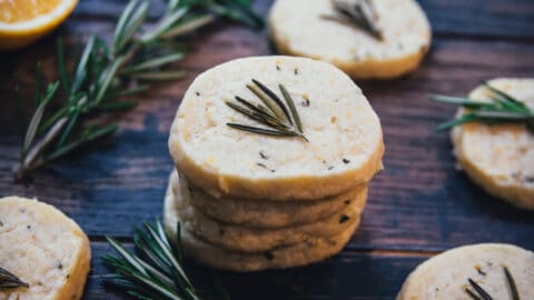 A stack of lemon rosemary shortbread cookies surrounded by single cookies each with a fresh rosemary frond on top, on a dark wood board scattered with rosemary and lemon halves.