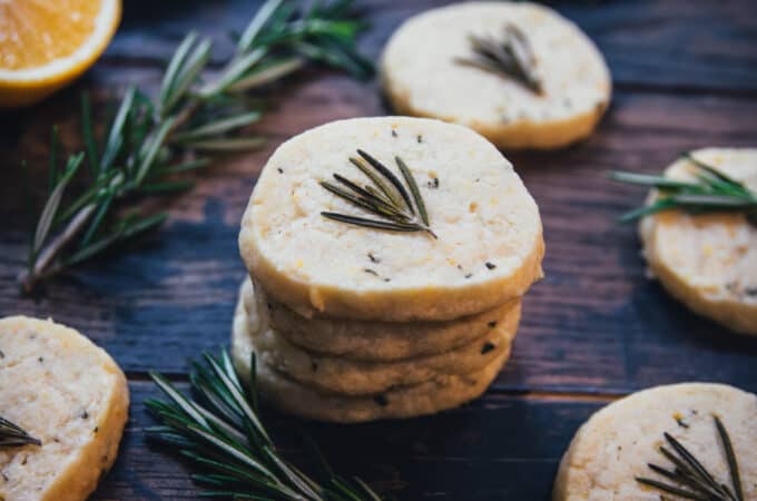 A stack of lemon rosemary shortbread cookies surrounded by single cookies each with a fresh rosemary frond on top, on a dark wood board scattered with rosemary and lemon halves.