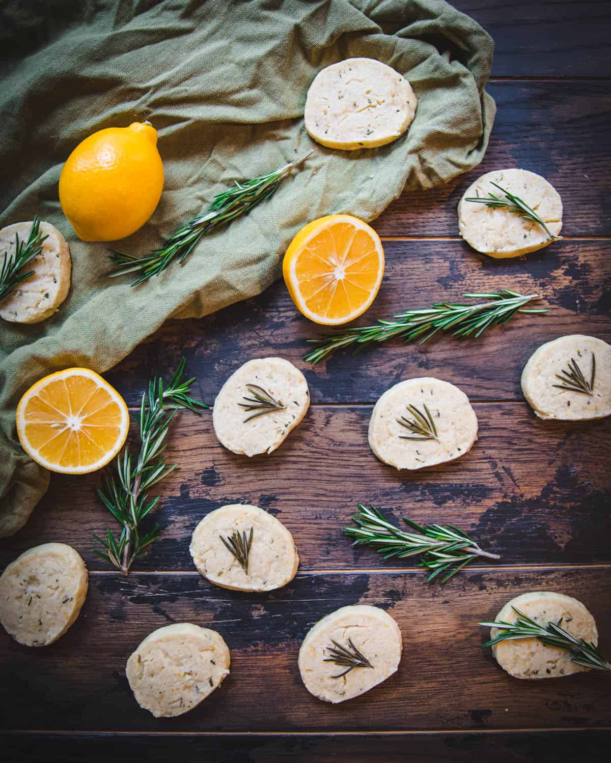 Lemon rosemary shortbread cookies on a dark wood surface with olive green fabric draped, and surrounded by fresh rosemary and lemon halves. 