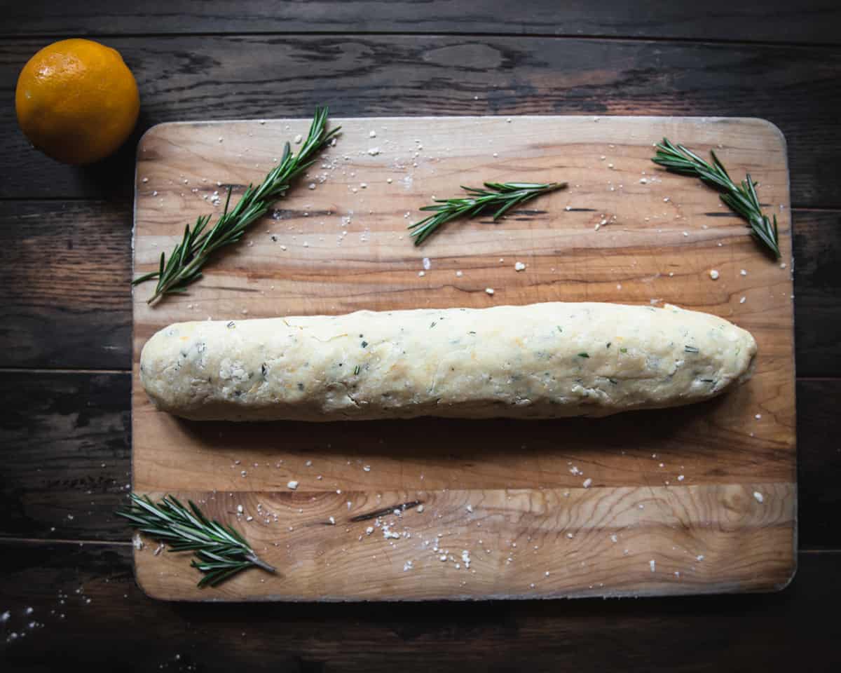 Lemon shortbread cookie dough formed into a log, on a wooden cutting board with fresh rosemary and a lemon surrounding.