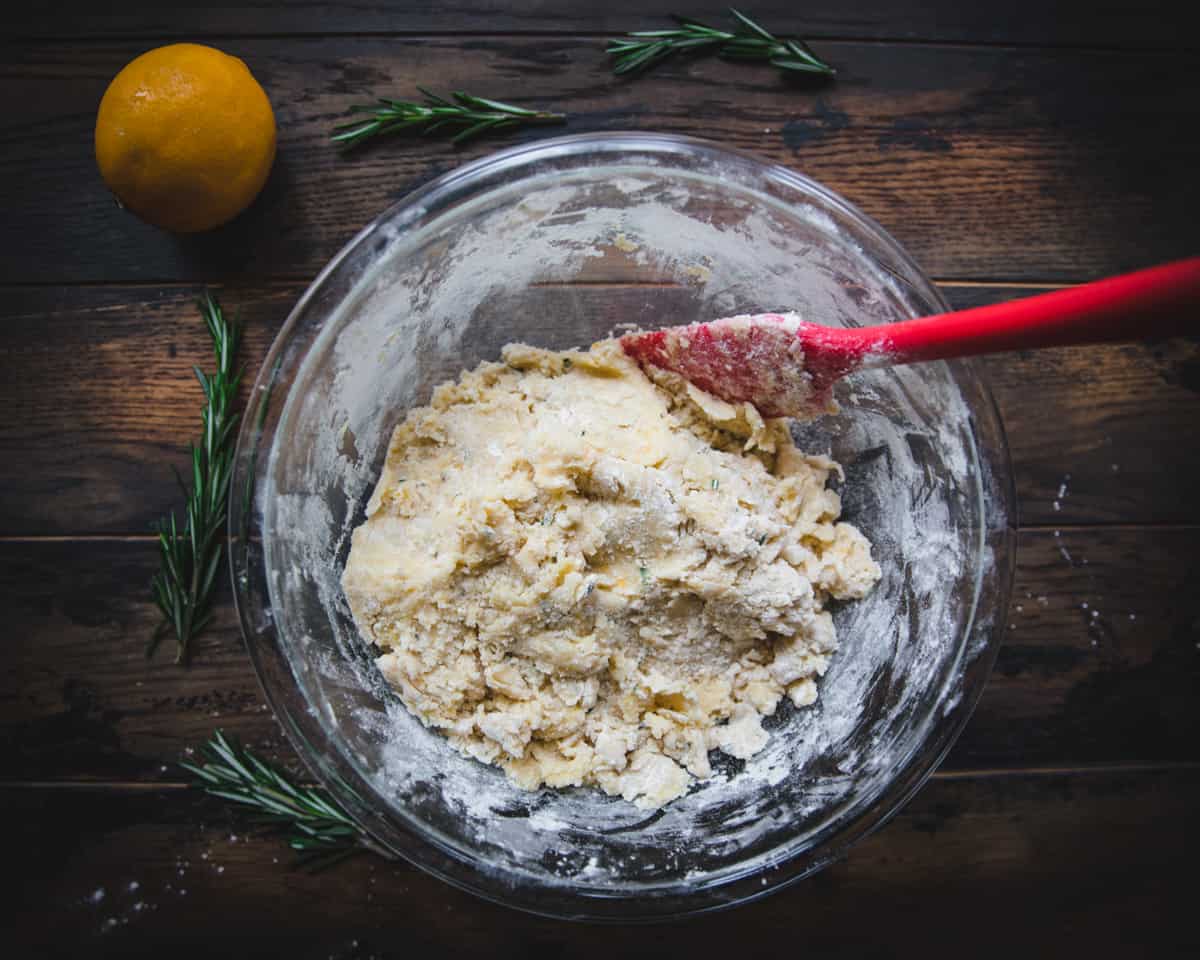 A dark wooden background with a clear bowl from top view with stirred lemon shortbread cookie dough with a red rubber spatula.