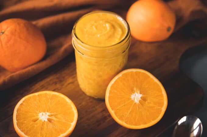 A jar of orange curd, with an orange sliced in half and whole oranges surrounding.