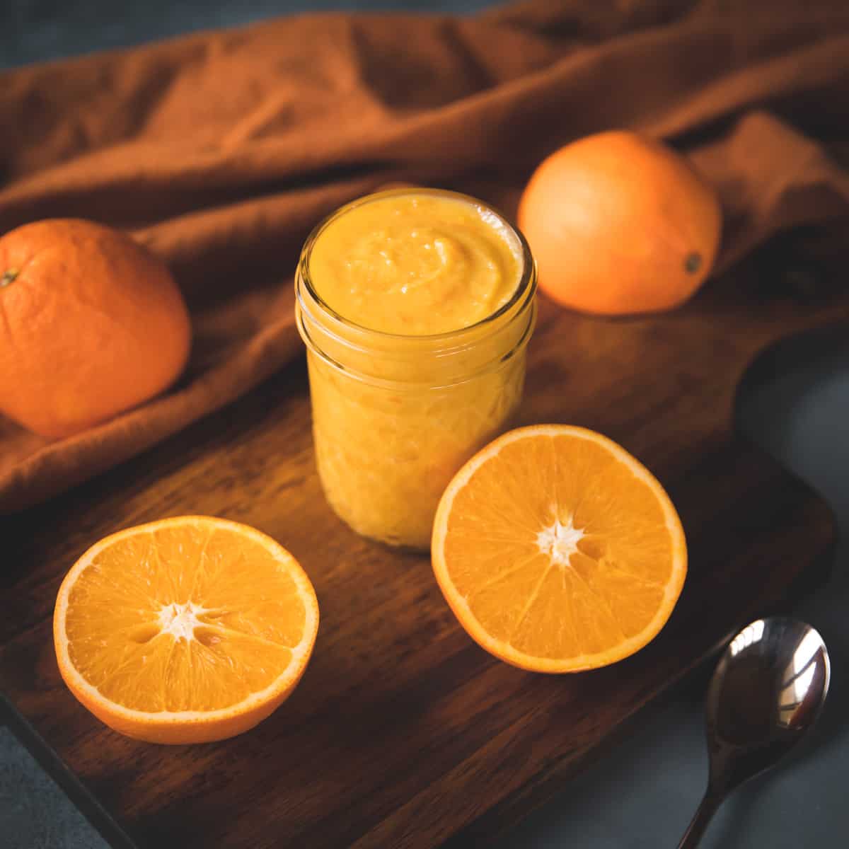 A jar of orange curd, with an orange sliced in half and whole oranges surrounding. 