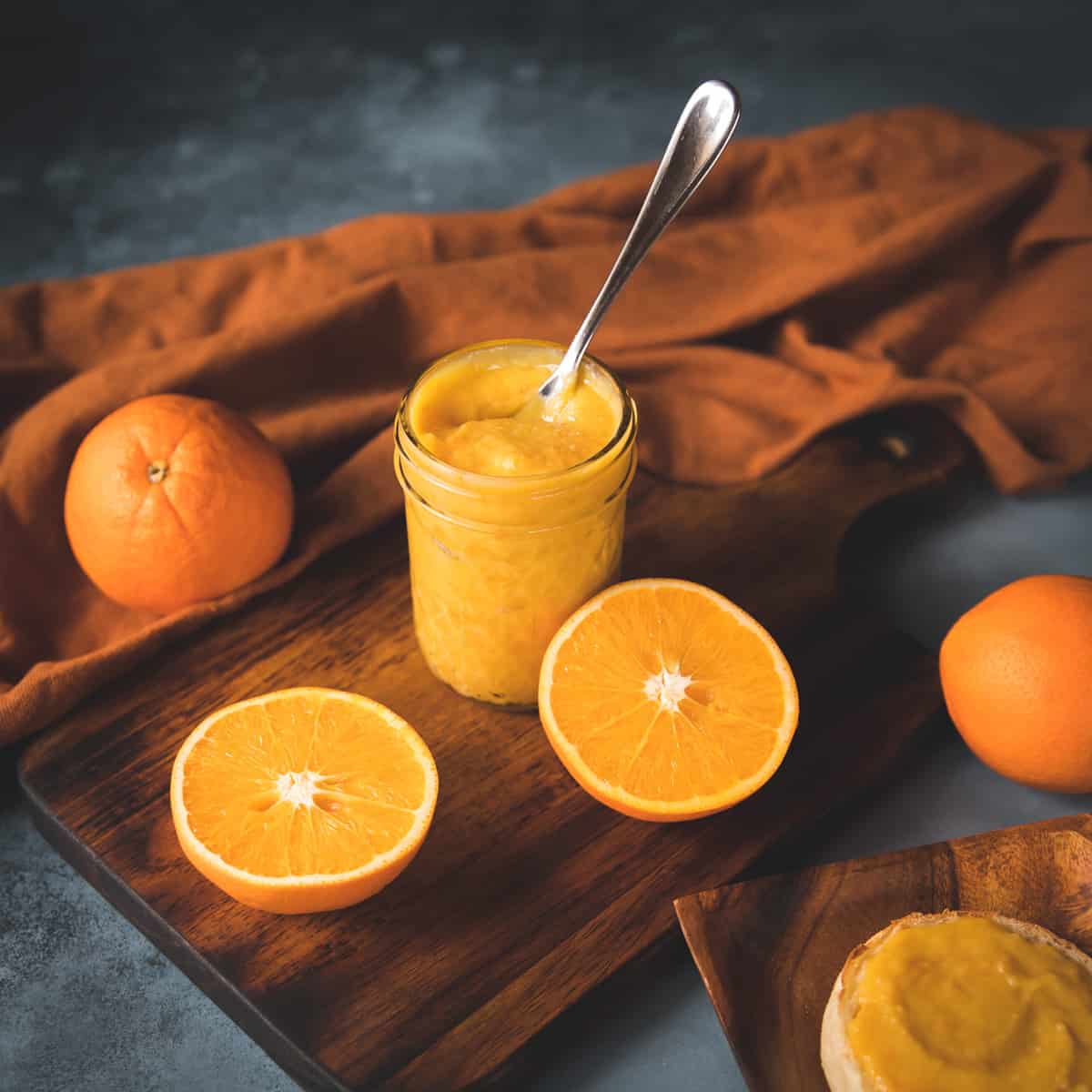 Orange curd in a jar with a spoon, on a wooden cutting board with halved oranges and whole oranges surrounding.