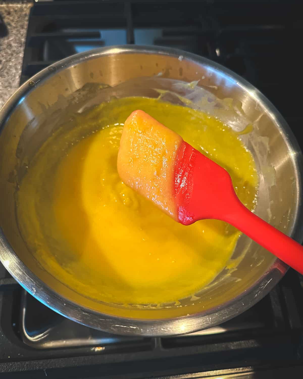 Orange curd on the spatula, showing it's thick enough to remain on the spatula, which is over the double boiler warming the thickening orange curd. 