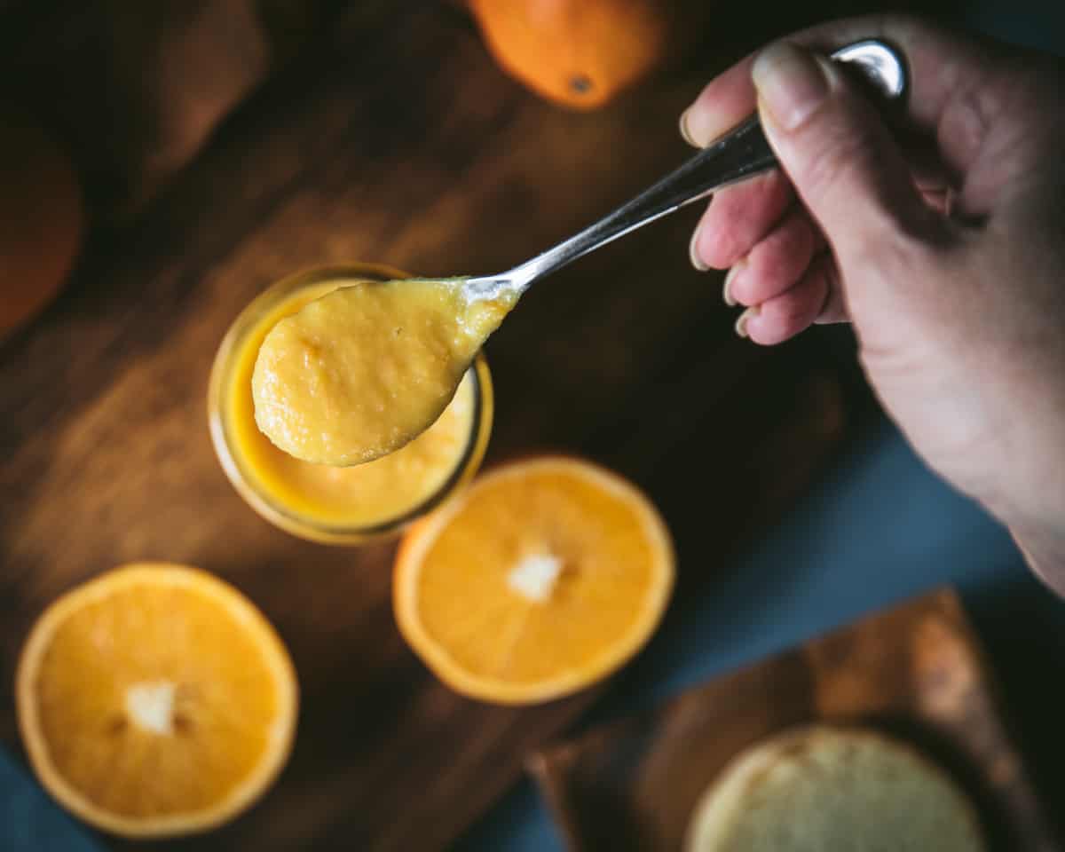 A jar of orange curd with a spoon filled with the curd lifted above, top view. Surrounded by oranges sliced in half face up.