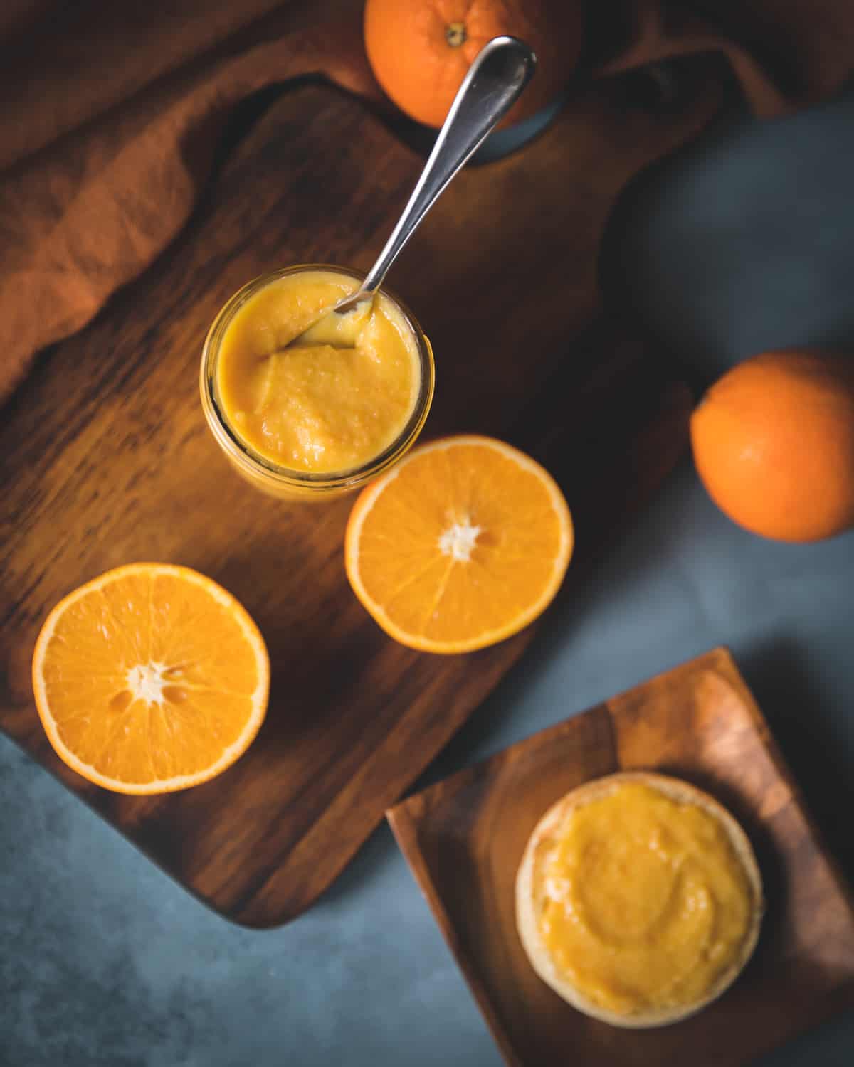 Top view of a jar of orange curd with a spoon, surrounded by halved and whole oranges on a wooden cutting board.