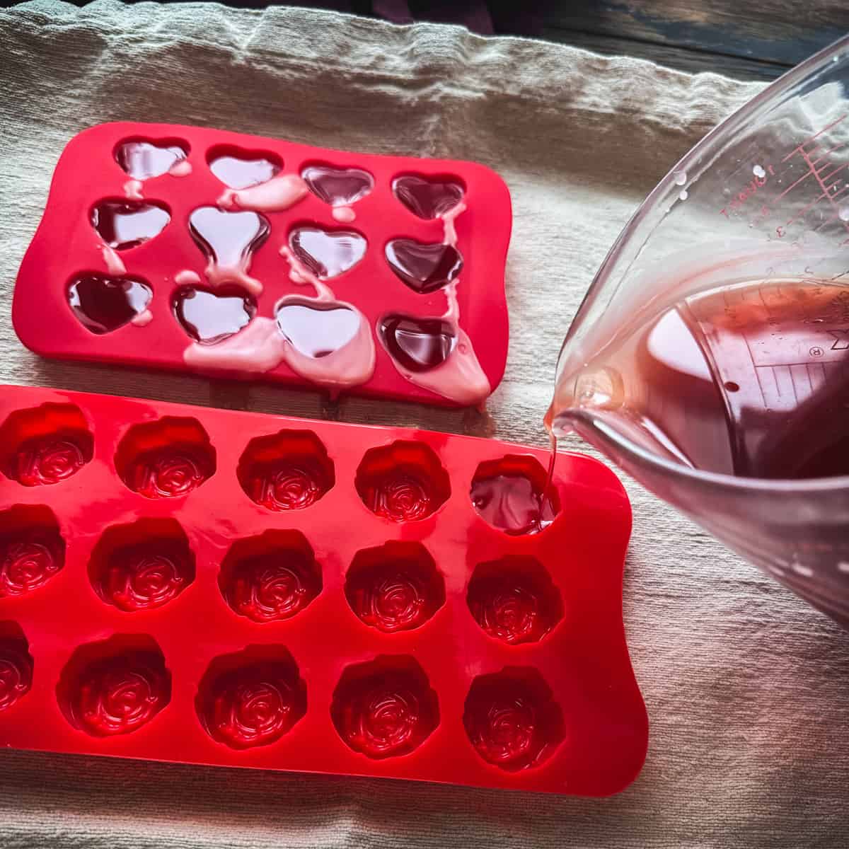 Rose infused oil melted mixture pouring into red heart shaped and flower shaped molds, sitting on a cloth.