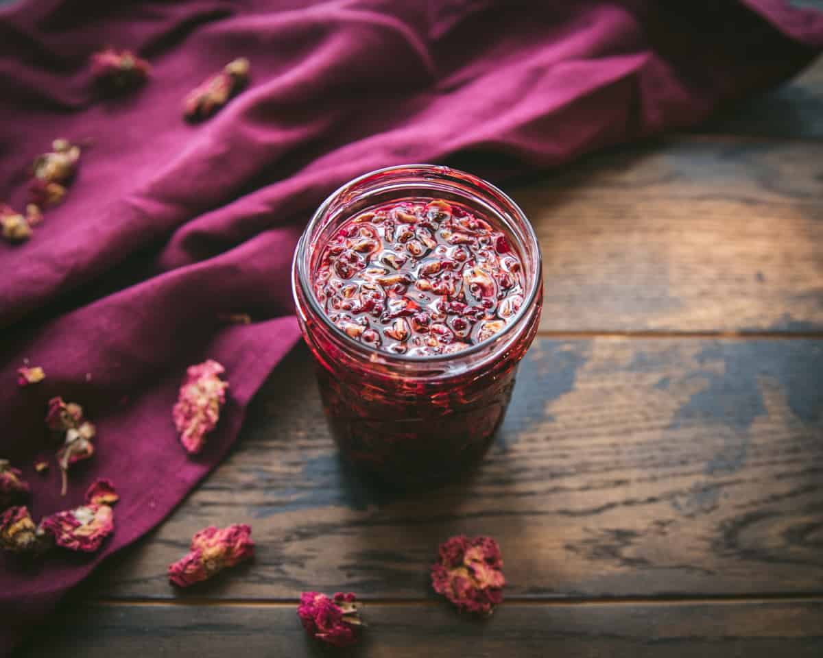 Infused rose petal and alkanet root in a jar that is red in color, on a dark wood and burgundy cloth surface sprinkled with dried rose petals.