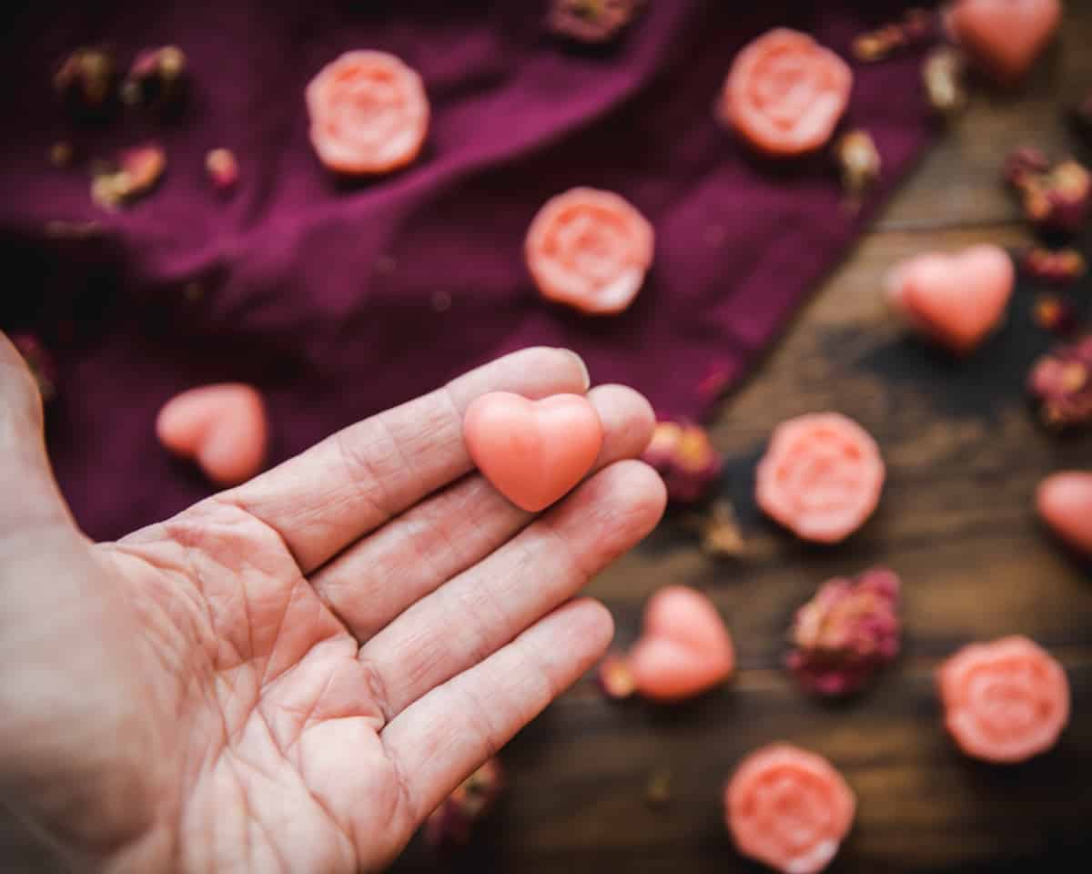 A hand holding up a heart lotion bar, in the background is scattered heart and flower shaped pink lotion bars on a dark wood surface with a burgundy cloth.