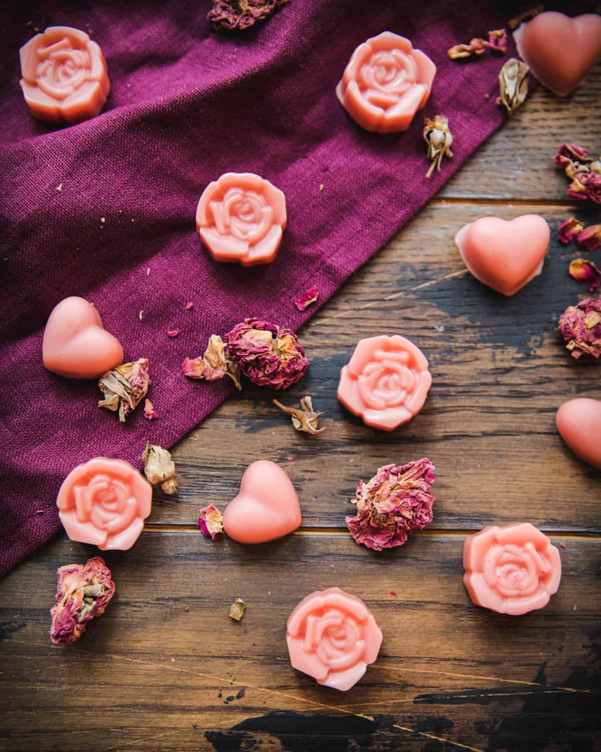 Rose and heart shaped pink lotion bars on a dark wood and burgundy cloth surface with scattered dried rose petals surrounding. 