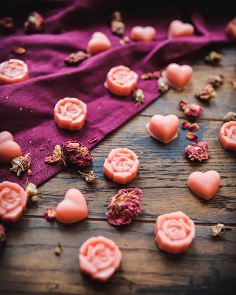 Rose and heart shaped pink lotion bars on a dark wood and burgundy cloth surface with scattered dried rose petals surrounding. 