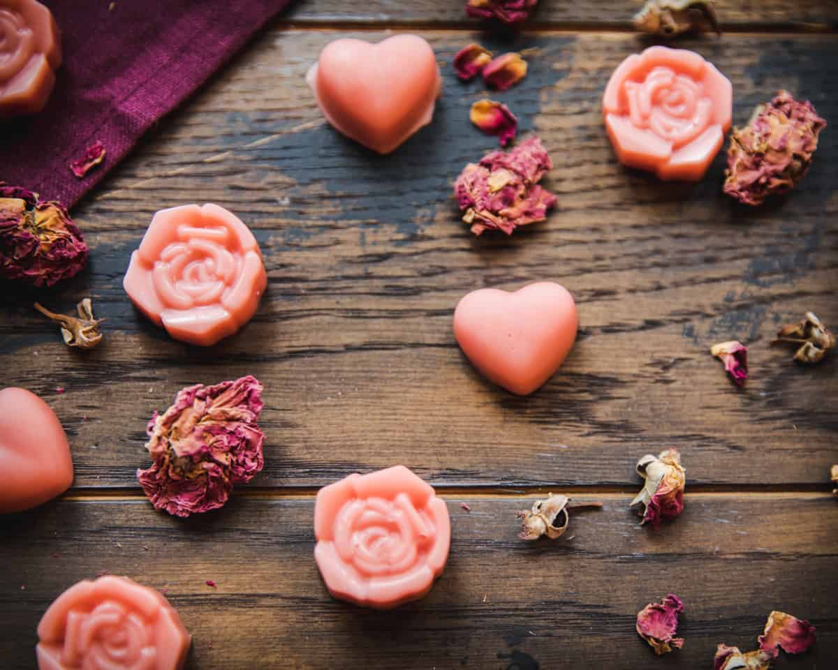 Close up of pink heart and rose shaped lotion bars surrounded by dried petals, on a dark wood surface.