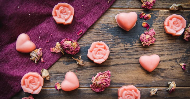 Rose shaped and heart shaped pink lotion bars, scattered with dried rose petals on a dark wood and burgundy cloth surface.