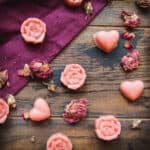 Pink lotion bars that are rose shaped and heart shaped scattered on a dark wood and burgundy cloth surface, with dried rose petals surrounding.