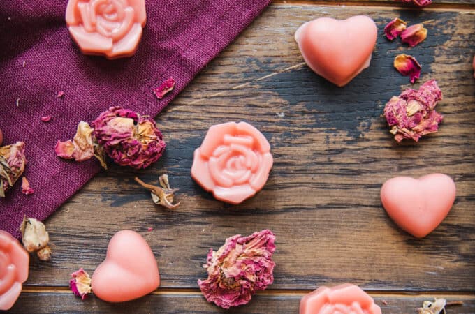 Pink lotion bars that are rose shaped and heart shaped scattered on a dark wood and burgundy cloth surface, with dried rose petals surrounding.