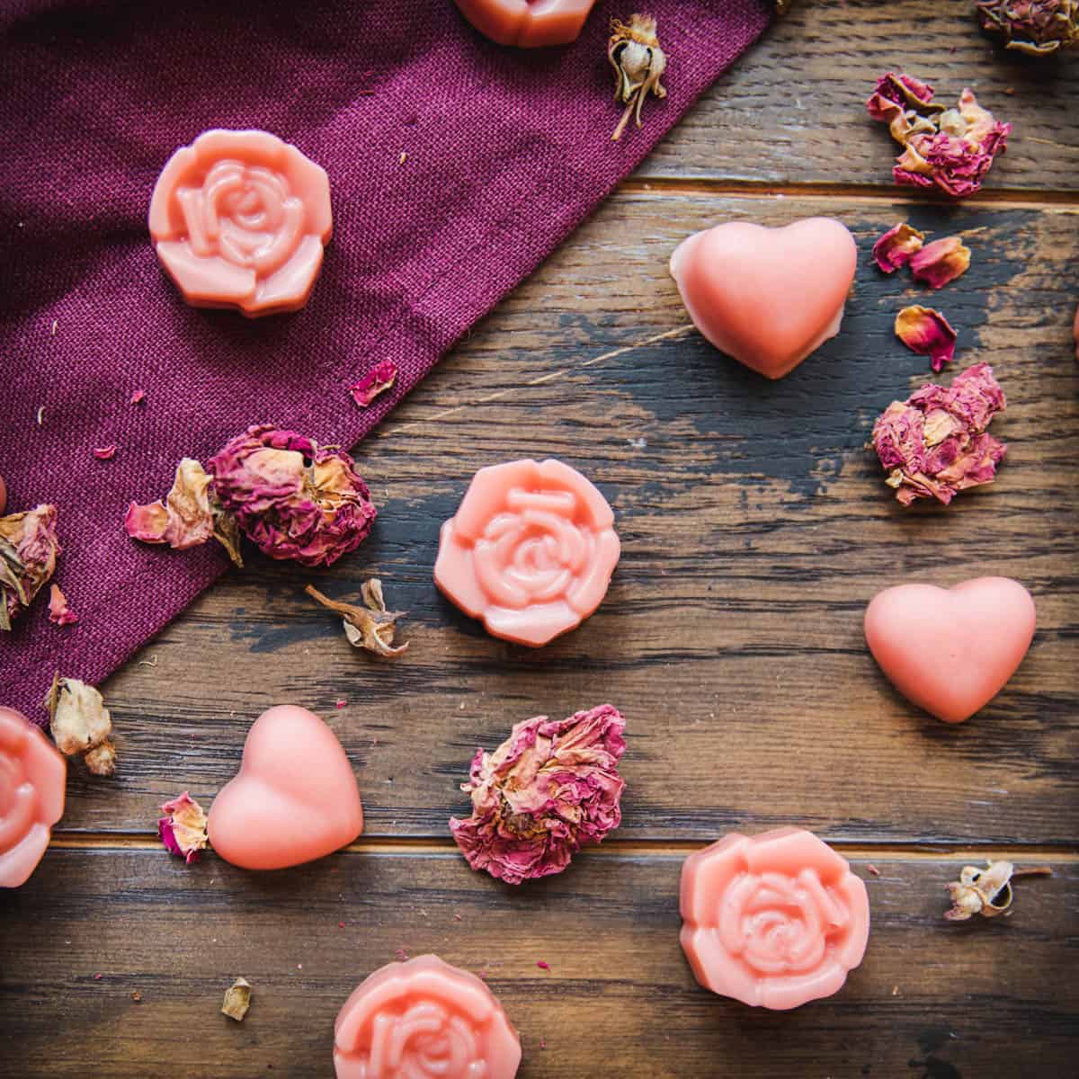 Pink lotion bars that are rose shaped and heart shaped scattered on a dark wood and burgundy cloth surface, with dried rose petals surrounding. 