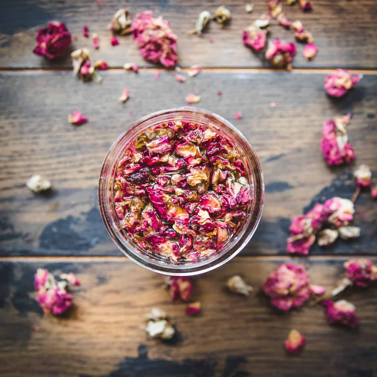 Top view of jar with dried rose petals infusing in oil, on a dark wood background scattered with dried rose petals. 