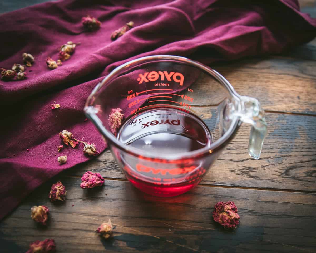 Rose infused oil that is a beautiful red color in a glass pour measuring cup, surrounded by dried rose petals on a dark wood and burgundy cloth surface. 