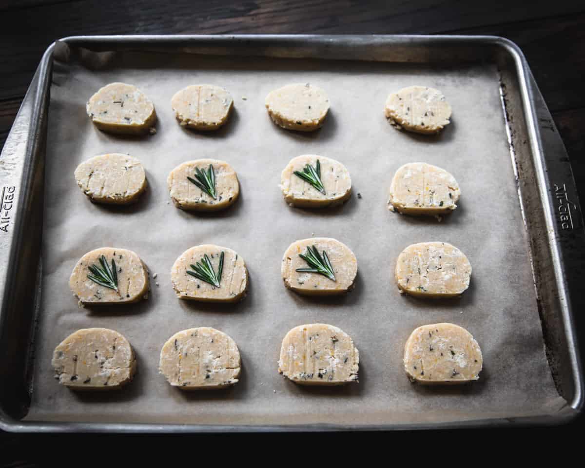 A parchment-lined baking sheet with unbaked lemon rosemary shortbread cookies, some topped with sprigs of rosemary. 