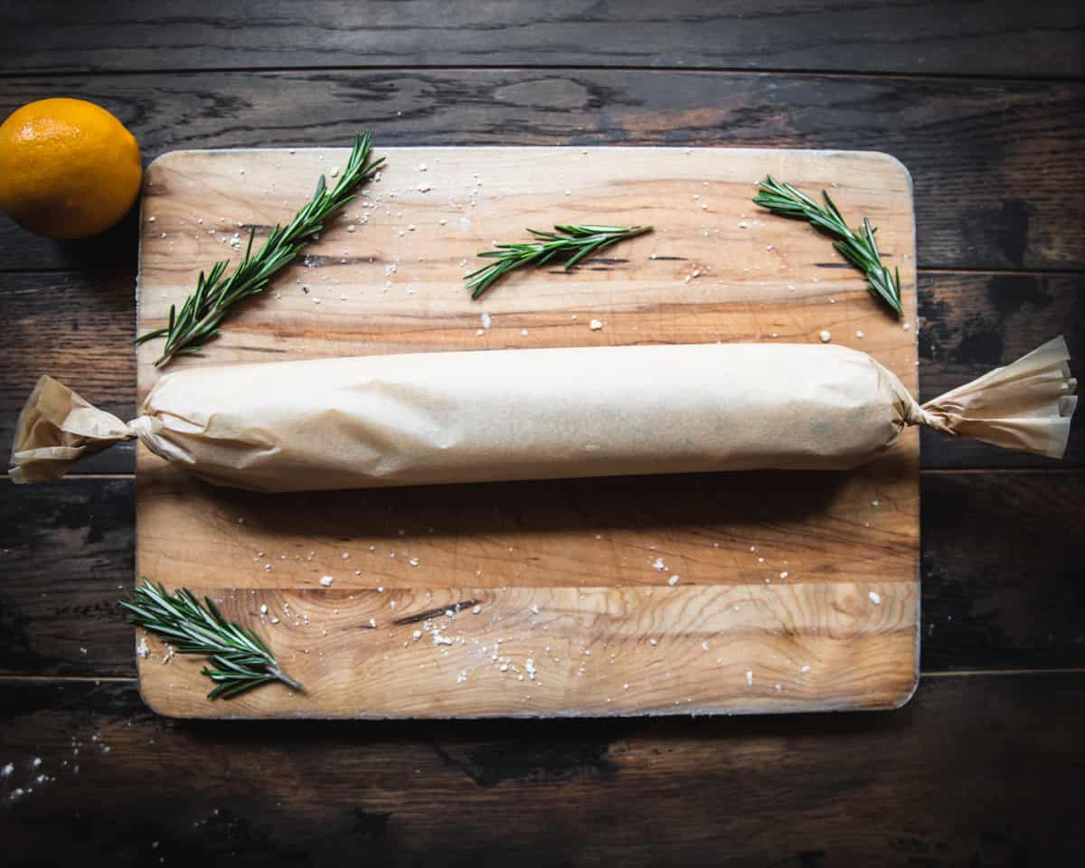 A dough log wrapped in parchment paper twisted on the ends on a wooden cutting board with fresh rosemary and a lemon surrounding. 