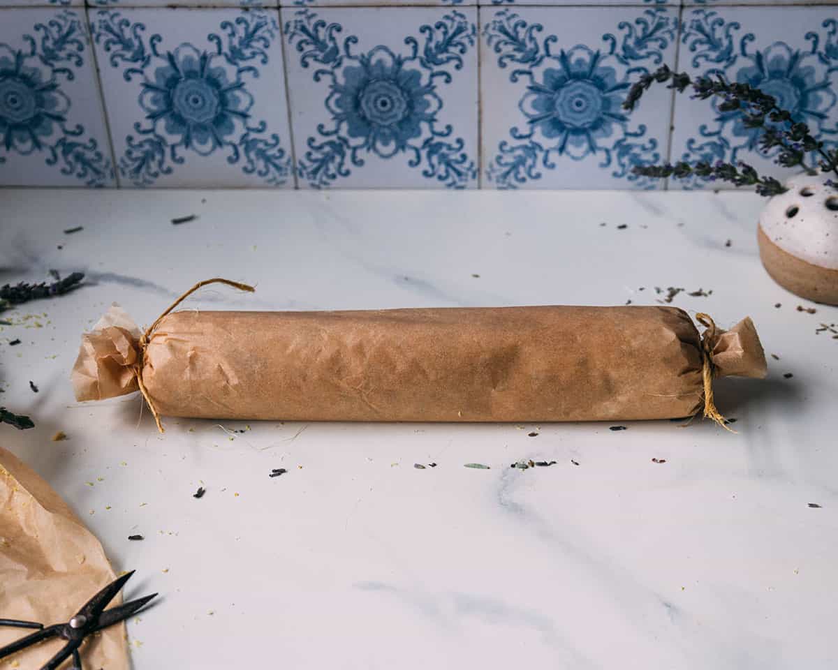 The dough log wrapped in parchment that is twisted at each end, on a white marble countertop with blue patterned tile in the background.