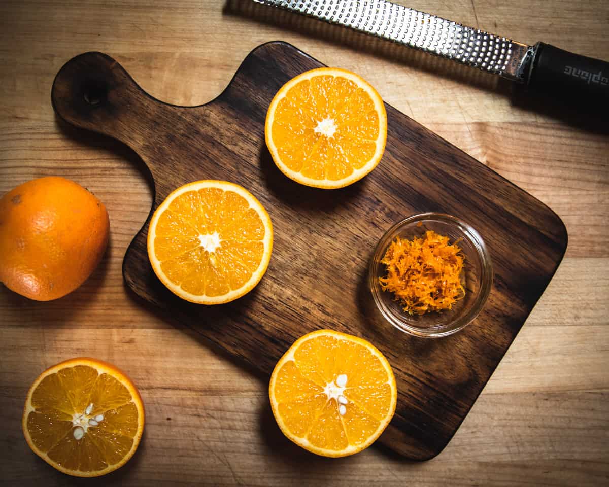 A small bowl of orange zest on a wooden cutting board surrounded by oranges cut in half face up. 