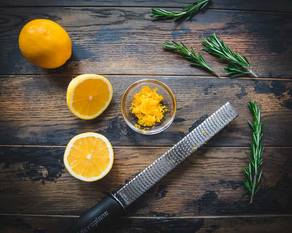 Dark wood with halved lemons, a bowl of lemon zest, a microplane, and fresh rosemary. 