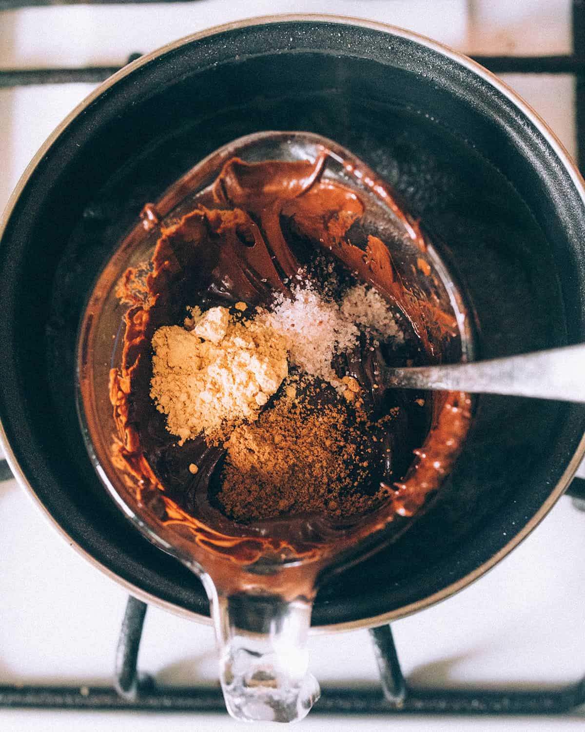 Mushroom powders in small piles on top of the melted chocolate in a makeshift double boiler, with a spoon ready to stir them in. 