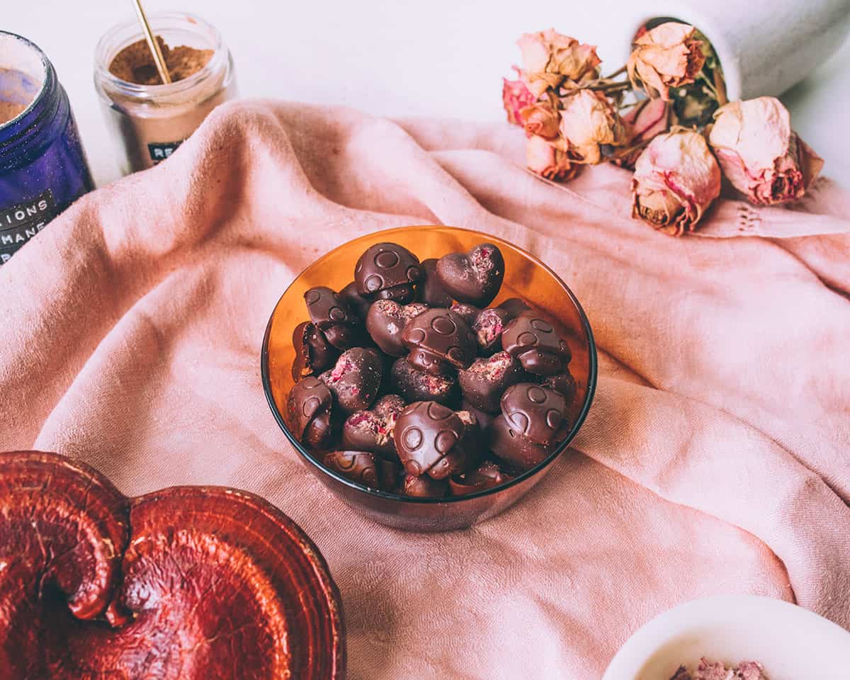 A small bowl of mushroom chocolates shaped like mushrooms and heart, on a surface draped with a pink cloth, surrounded by a big reishi mushroom, and pink roses.