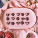 A pink oval plate with little heart and mushroom shaped chocolates lined up in 3 rows, surrounded by pink fabric and pink roses.