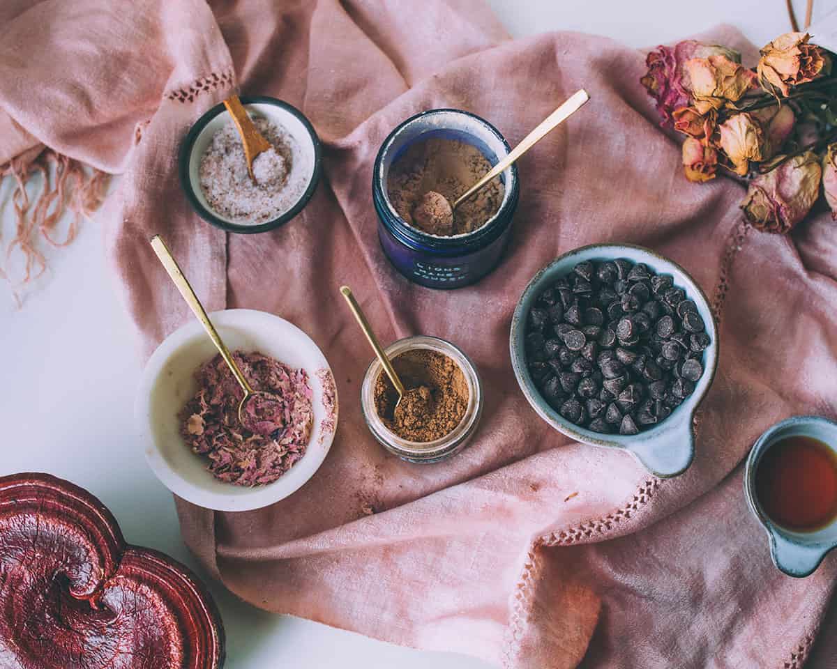 Bowls of ingredients with gold spoons in them, on a white surface with a ping cloth draped. 