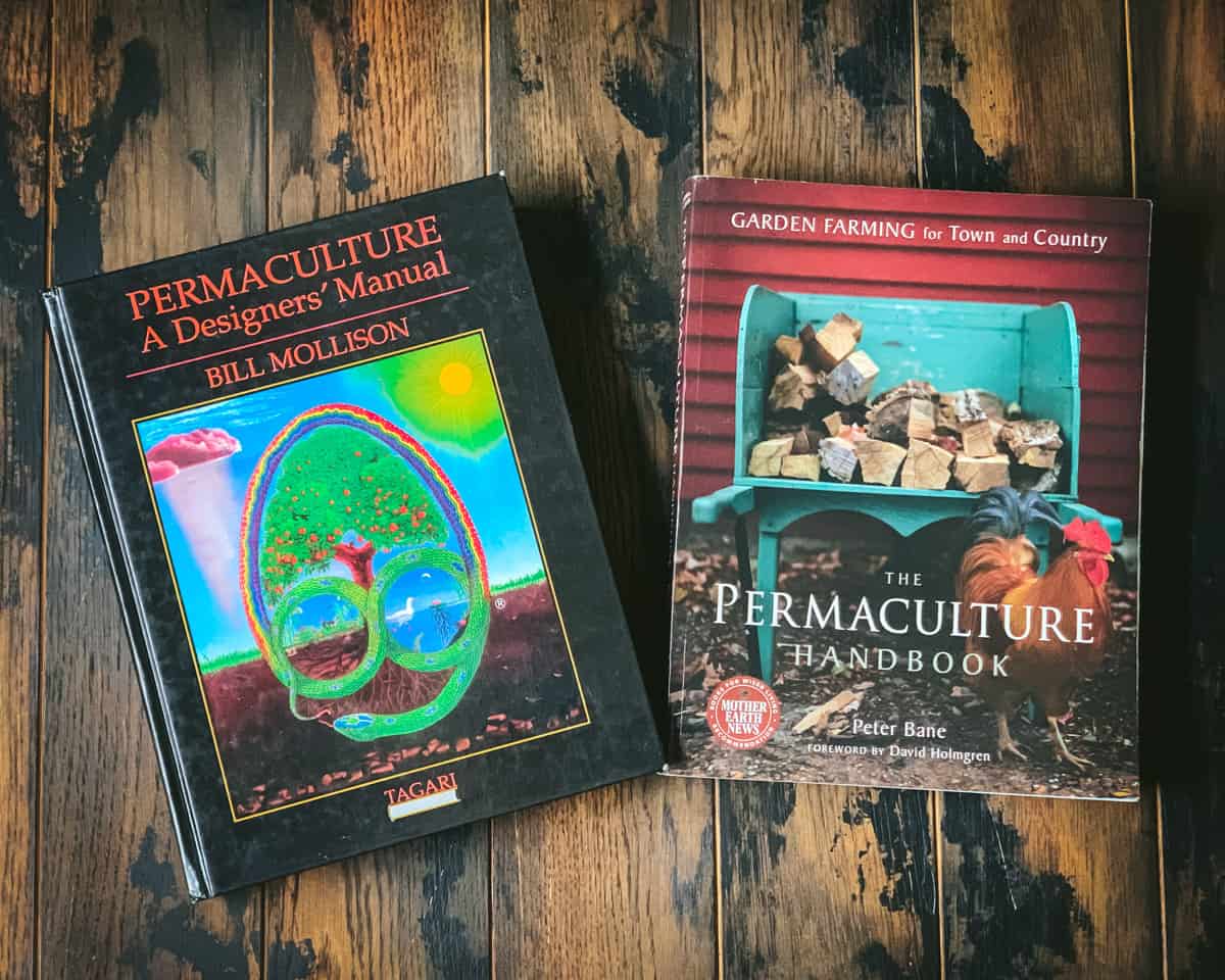 2 permaculture books lying side by side on a dark wood surface. 