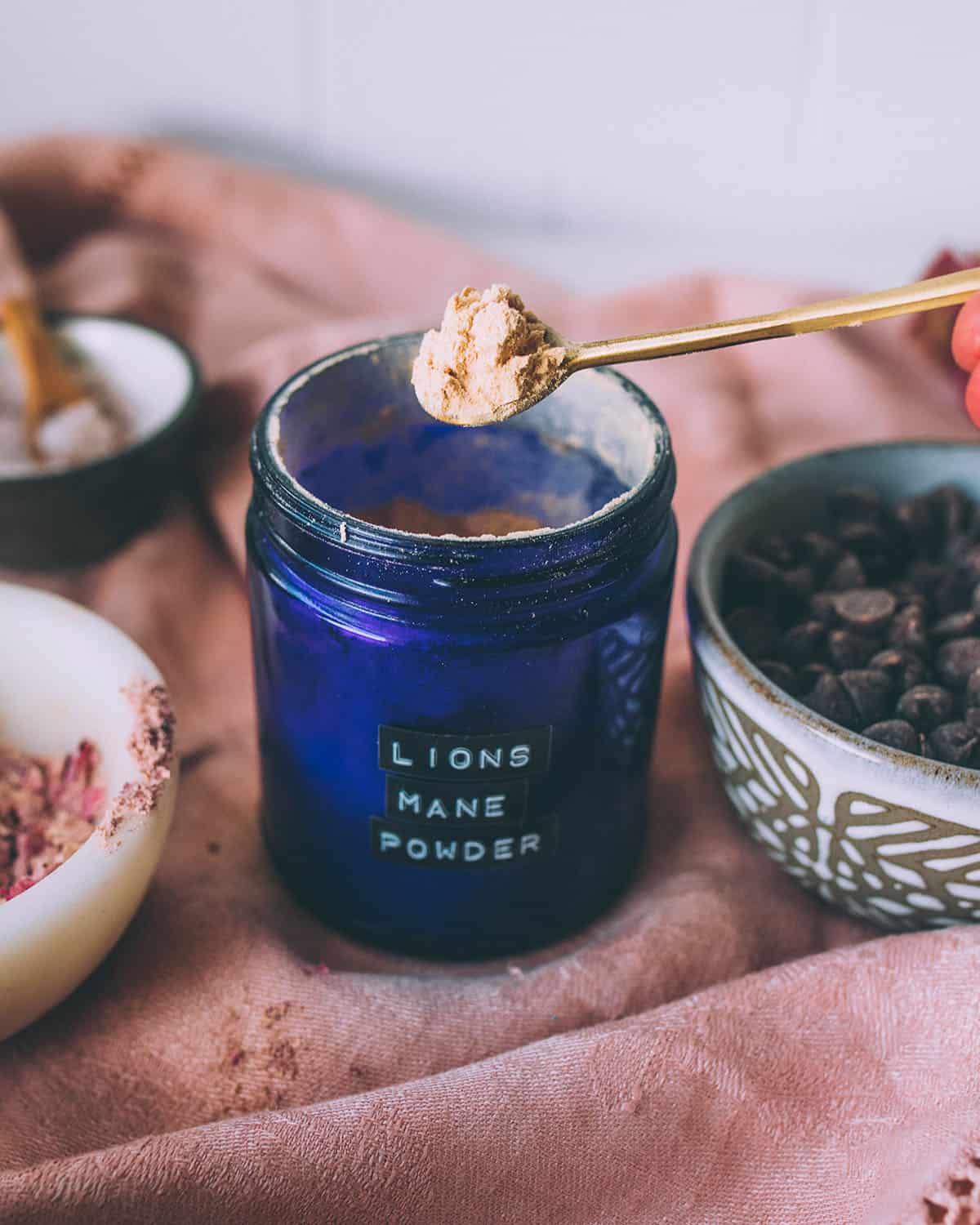 A blue jar labelled "lion's made powder" with a gold spoon heaping with the powder over the top, on a surface with a pink cloth and other ingredients partially seen in bowls surrounding.