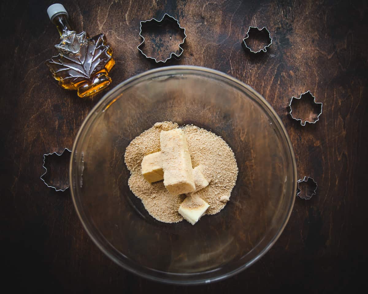 A clear bowl with butter and tan colored maple sugar, on a dark wood surface surrounded by a glass maple leaf shaped syrup and maple leaf shaped cookie cutters. 