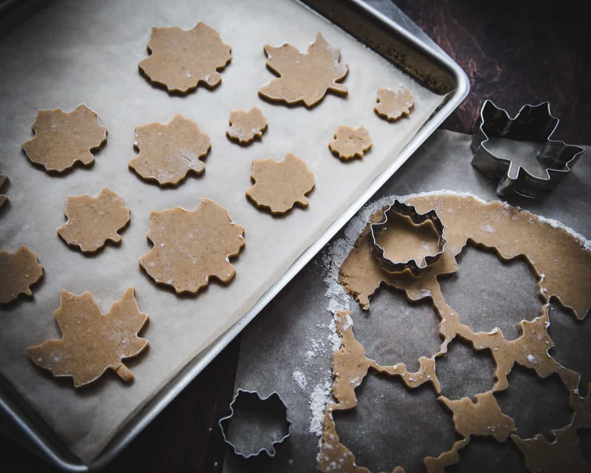 A sheet pan with cut out maple leaf dough, also showing dough rolled out on parchment being cut with maple leaf cookie cutters. 