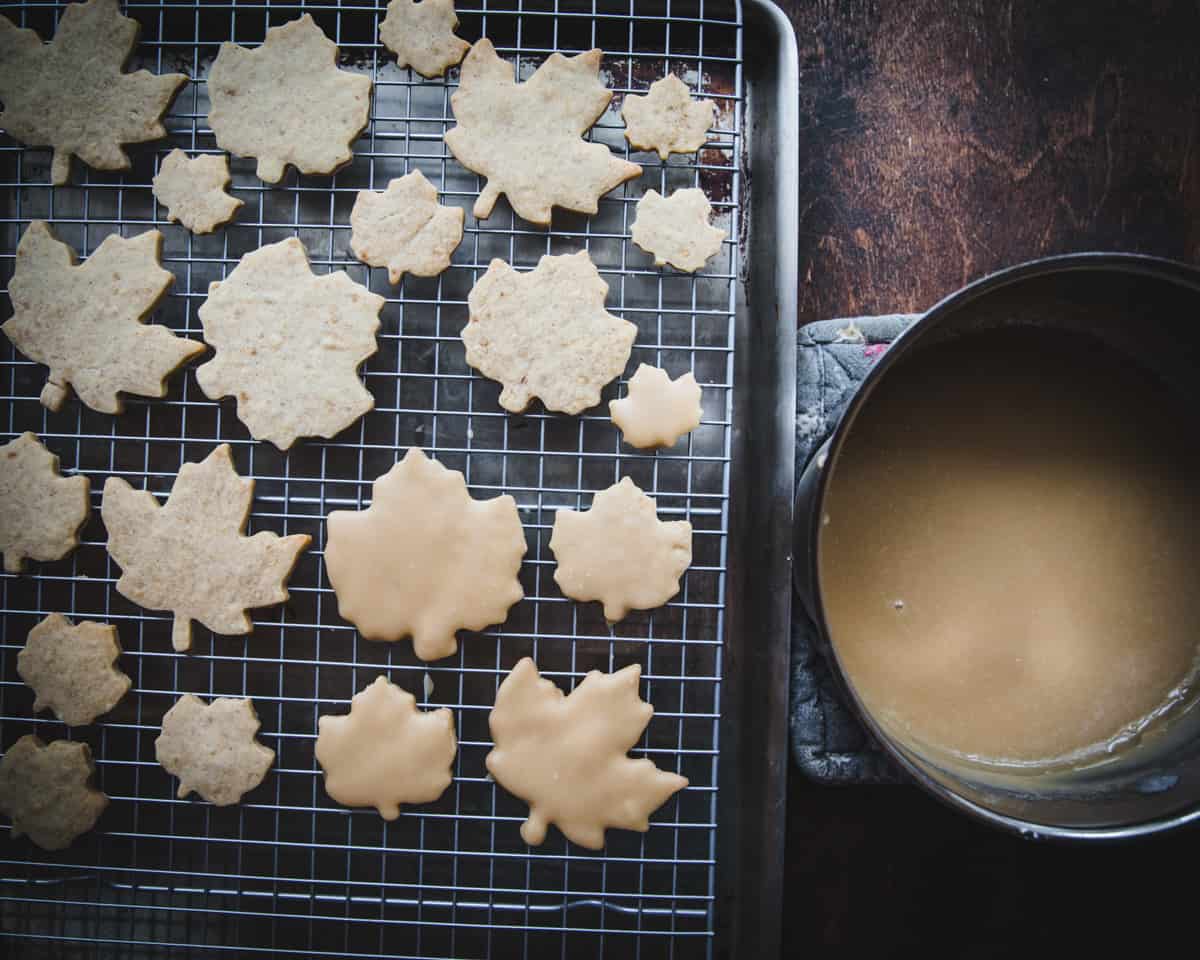 A pot of maple cream icing next to a cooling rack on a baking sheet, with some cookies iced and some not yet iced. 