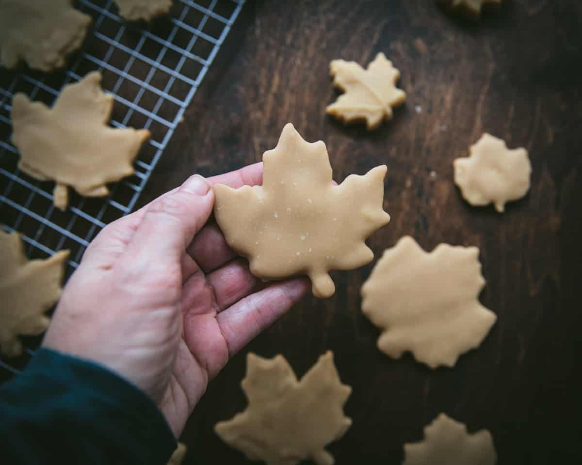 A hand holding up a maple leaf cookie with maple icing, below is more cookies and a cooling rack on a dark wood surface. 