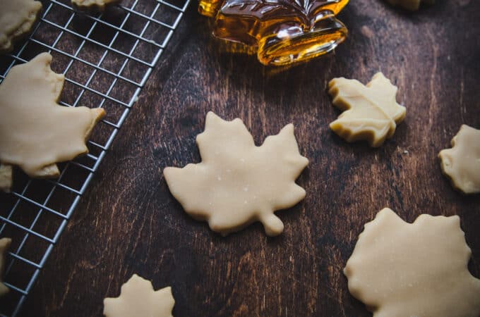 Maple leaf shaped cookies of different sizes on a dark wood surface, some on a cooling rack, along with maple syrup in a maple shaped glass container.