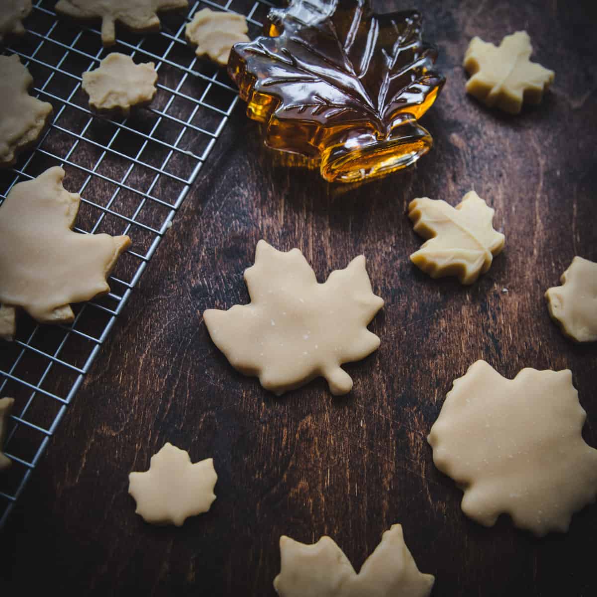 Maple leaf shaped cookies of different sizes on a dark wood surface, some on a cooling rack, along with maple syrup in a maple shaped glass container. 