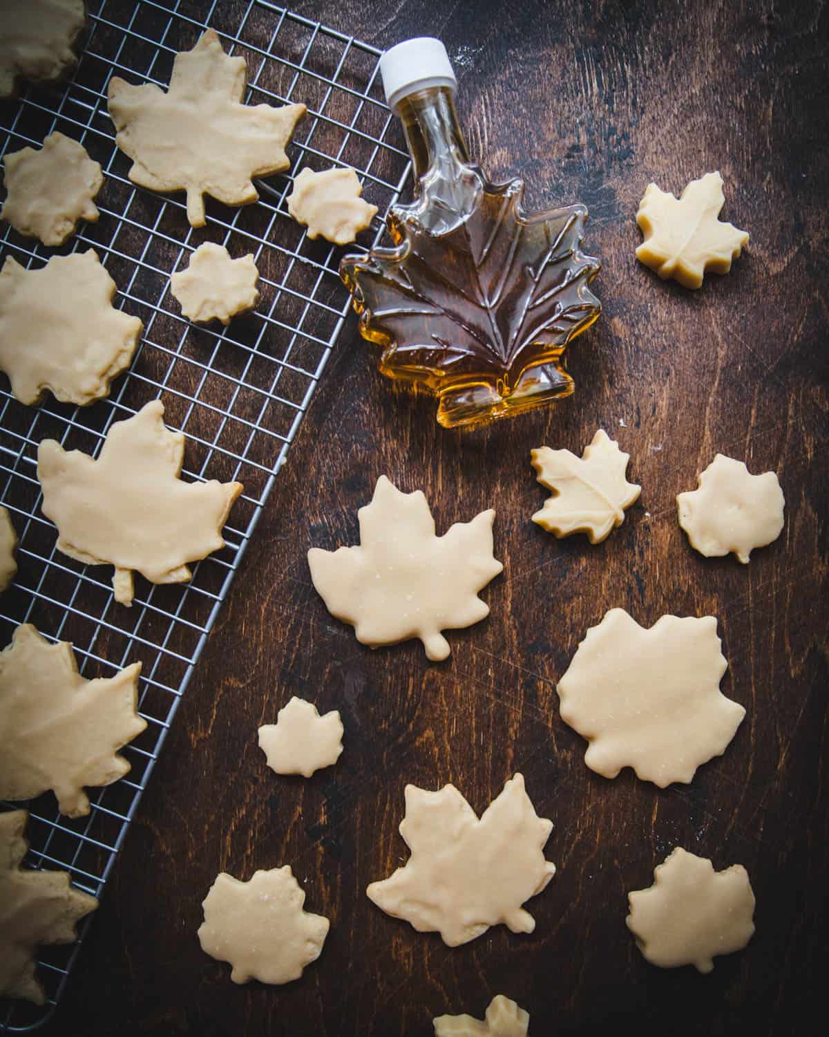 Maple leaf cookies on a dark wood surface and a cooling rack, with a jar of maple leaf shaped syrup. 